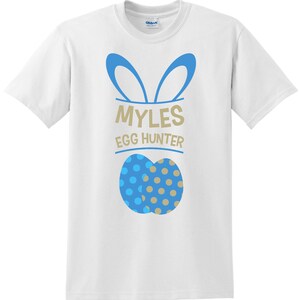 May include: White t-shirt with a blue and gold Easter egg design. The shirt features the text "MYLES EGG HUNTER" in gold and blue. The design includes blue bunny ears above the text.