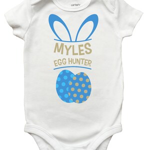 May include: White baby onesie with blue bunny ears and the text "MYLES EGG HUNTER". Two blue and tan polka-dotted Easter eggs are printed below the text. The onesie is a Carter's brand.