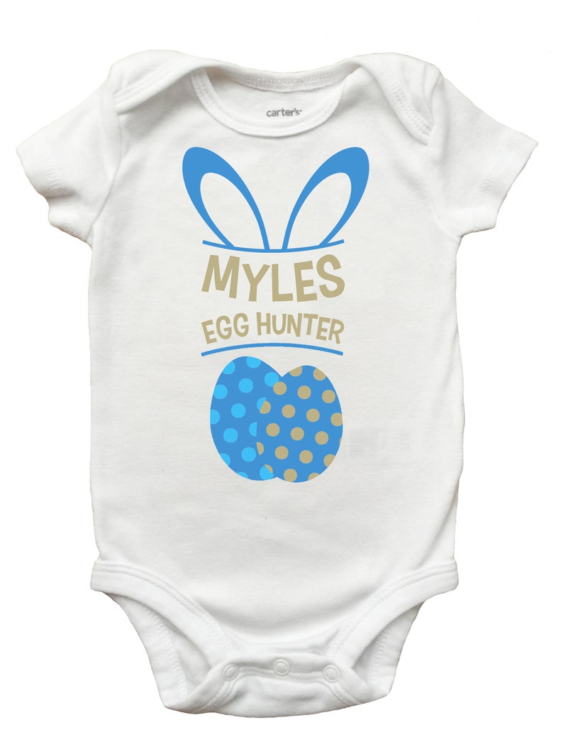May include: White baby onesie with blue bunny ears and the text "MYLES EGG HUNTER". Two blue and tan polka-dotted Easter eggs are printed below the text. The onesie is a Carter's brand.