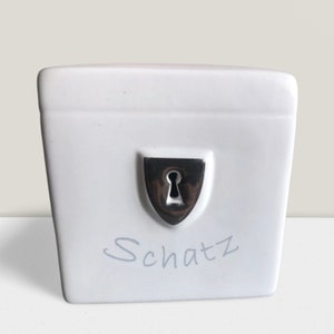 Può includere: Scatola in ceramica bianca con un design a forma di buco della serratura argentato e la parola "Schatz" scritta in grigio.
