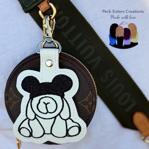 May include: A brown and white round coin purse with a cartoon bear design. The purse has a gold zipper and a white leather strap with a silver clasp. The strap is attached to a green strap with the words "LOUIS VUITTON" printed on it.