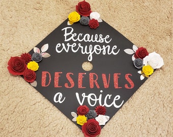 Puede incluir: Un adorno para birrete de graduación negro decorado con flores de papel en rojo, amarillo, blanco y gris. El texto "Because everyone DESERVES a voice" aparece en letras de purpurina blancas y rojas sobre la superficie cuadrada.