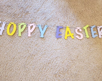 Pode incluir: Uma faixa decorativa com letras recortadas individuais que formam a frase HOPPY EASTER. Cada letra é feita em uma cor pastel diferente, incluindo amarelo, rosa, azul, roxo e verde claro, todas presas em um barbante fino.