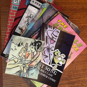 May include: A collection of comic books with various covers. The covers feature illustrations and text, including "CR," "A ROLE," "ST SONG," and "BOLT CASE." The color palette includes red, pink, blue, and black.