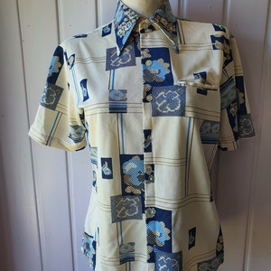 Puede incluir: Una camisa de manga corta con botones y un patrón geométrico en tonos de azul, marrón y crema. La camisa tiene un cuello puntiagudo y un solo bolsillo en el pecho.
