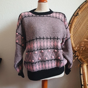 May include: A long-sleeve knit sweater featuring a black and pink argyle-inspired pattern with small raised pom-pom details. The garment has a black ribbed crew neckline, black ribbed cuffs, and a black ribbed hem, constructed with a textured knit.