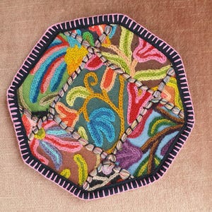 May include: A colorful, embroidered textile with a geometric shape. The design features a variety of floral and abstract patterns in shades of blue, green, pink, orange, and yellow. The edges are outlined with a black border and pink stitching.