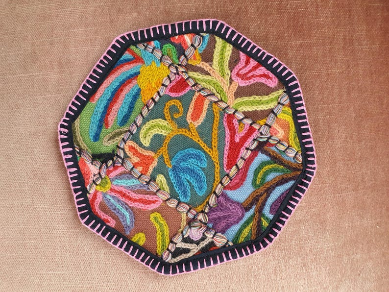 May include: A colorful, embroidered textile with a geometric shape. The design features a variety of floral and abstract patterns in shades of blue, green, pink, orange, and yellow. The edges are outlined with a black border and pink stitching.