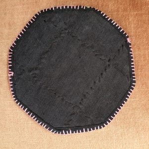 May include: Black, octagonal coaster with pink stitching around the edge. The coaster has a textured surface with visible stitching across the center. The background is a solid, light brown color.