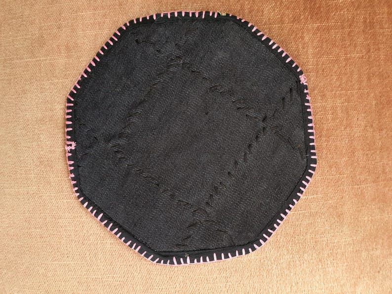 May include: Black, octagonal coaster with pink stitching around the edge. The coaster has a textured surface with visible stitching across the center. The background is a solid, light brown color.