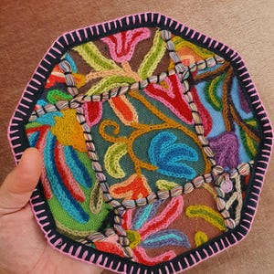 May include: A colorful, embroidered textile with a geometric shape. The design features a variety of floral and abstract patterns in shades of red, blue, green, and orange, outlined with black and pink stitching. The textile is held by a hand.