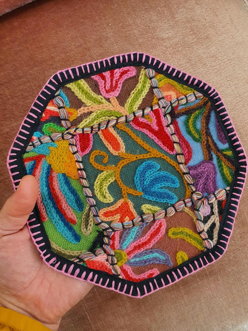 May include: A colorful, embroidered textile with a geometric shape. The design features a variety of floral and abstract patterns in shades of red, blue, green, and orange, outlined with black and pink stitching. The textile is held by a hand.