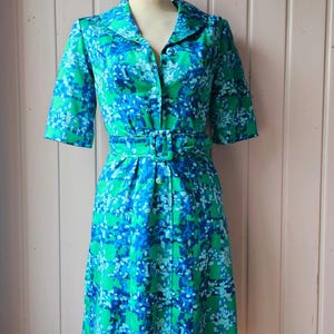 Könnte beinhalten: Ein Vintage-Kleid mit grünem und blauem Blumendruck, Knopfleiste und Gürtel. Das Kleid hat kurze Ärmel und einen knielangen Rock.