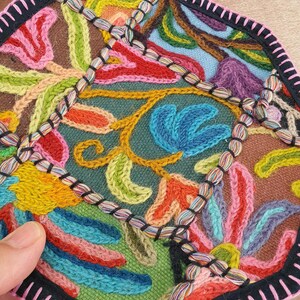 May include: Close-up of a colorful, embroidered textile with a floral design. The piece features a variety of colors including blue, red, yellow, and green. The edges are finished with a black border and pink trim. The embroidery is detailed and intricate.