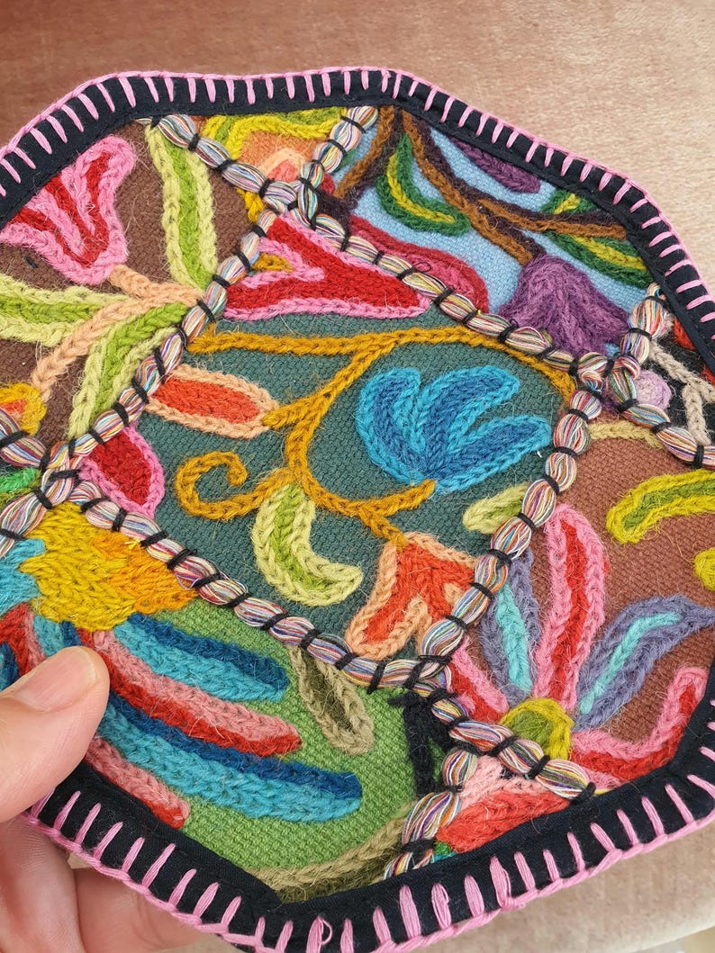 May include: Close-up of a colorful, embroidered textile with a floral design. The piece features a variety of colors including blue, red, yellow, and green. The edges are finished with a black border and pink trim. The embroidery is detailed and intricate.