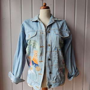 Puede incluir: Una chaqueta vaquera azul claro con osos y flores bordadas. Los osos llevan ropa colorida y están sentados en una playa.