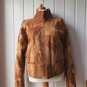 Peut inclure: Veste en daim marron avec un motif patchwork. La veste a un col montant, une fermeture éclair sur toute la longueur et une petite poche zippée sur la poitrine. La veste est présentée sur un mannequin.