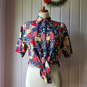 Op de afbeelding: Een button-down shirt met korte mouwen op een mannequin. Het shirt heeft een patchwork-ontwerp met rode, gele en witte bloemen- en geometrische patronen op een marineblauwe achtergrond. Het shirt is in de taille geknoopt.