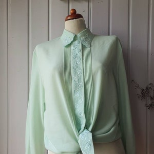 Pode incluir: Uma blusa verde claro de manga comprida com gola e bordado com motivos de folhas. A blusa tem pregas na frente e é amarrada na cintura. A blusa é exibida em um manequim.