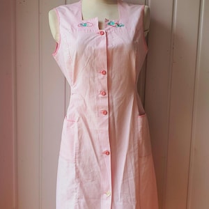 May include: A vintage pink and white striped apron dress with a square neckline and button-down front. The dress features two front pockets and embroidered floral details near the neckline. The dress is displayed on a mannequin.