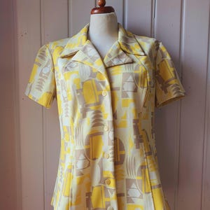 May include: A vintage yellow button-down shirt with a geometric pattern in shades of yellow, beige, and gray. The short-sleeved shirt is displayed on a mannequin. The shirt has a collared neckline and a series of buttons down the front.