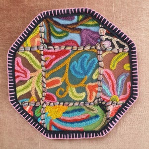 May include: An octagonal, embroidered coaster with a colorful floral design. The coaster features a black border with pink stitching and is filled with vibrant hues of blue, green, orange, and purple. The background is a muted brown.