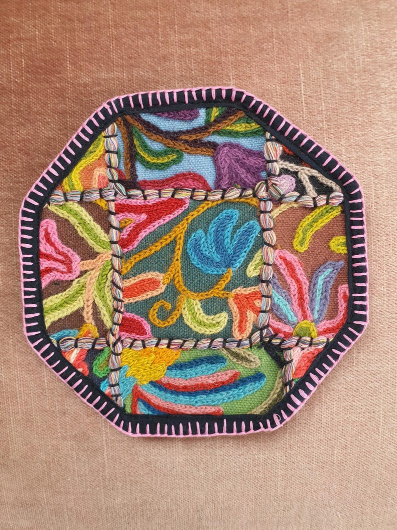May include: An octagonal, embroidered coaster with a colorful floral design. The coaster features a black border with pink stitching and is filled with vibrant hues of blue, green, orange, and purple. The background is a muted brown.