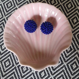 May include: A pair of blue beaded stud earrings rest in a pink seashell-shaped dish. The earrings are round and covered in small, dark blue beads. The dish is light pink and sits on a black and white patterned surface.