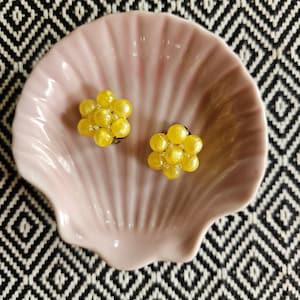 Könnte beinhalten: Ein Paar gelbe, perlenbesetzte Cluster-Ohrringe, präsentiert auf einer rosa, muschelförmigen Schale. Die Ohrringe bestehen aus kleinen, runden Perlen, die zu Clustern angeordnet sind. Die Schale liegt auf einer schwarz-weiß gemusterten Oberfläche.