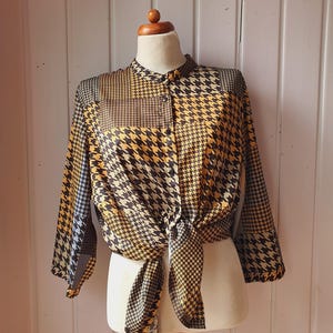 Könnte beinhalten: Eine Bluse mit Hahnentrittmuster in Schwarz, Gold und Weiß, die an der Taille gebunden ist. Das langärmlige Oberteil wird auf einer Schaufensterpuppe präsentiert. Die Bluse hat einen Stehkragen und eine Knopfleiste.