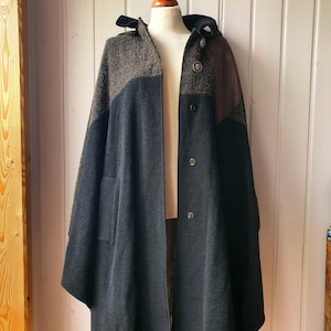 May include: A dark grey and brown hooded cape with a button-up front. The cape features a textured, patterned upper section and a solid, flowing lower section. The cape is displayed on a mannequin, showcasing its design and drape.