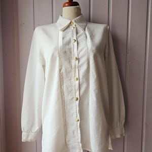 Pode incluir: Uma blusa branca de manga comprida com gola e botões dourados. A frente apresenta um painel texturizado com um design geométrico. A blusa é exibida em um manequim de madeira.