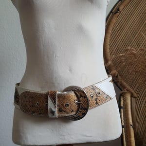 May include: A wide belt featuring a snakeskin pattern in shades of brown and tan, accented with white panels. The belt includes a large, round, metallic buckle with intricate engravings and silver-toned metal eyelets along the strap length.