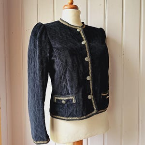 Puede incluir: Chaqueta vintage de terciopelo negro con mangas acolchadas y corte entallado. La chaqueta presenta ribetes dorados a lo largo del cuello, los bolsillos y los cierres de botones. La chaqueta se exhibe en un maniquí.