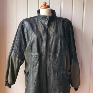 Könnte beinhalten: Eine dunkel anthrazitfarbene Lederjacke mit Stehkragen und einem einzelnen Druckknopfverschluss, langen Ärmeln mit gerippten Bündchen und zwei Vordertaschen. Die Jacke weist ein strukturiertes, blattartiges Muster auf den Lederpaneelen auf.
