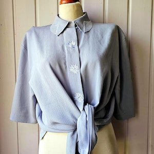 Pode incluir: Uma blusa azul clara com mangas curtas, exibida em um manequim. A blusa apresenta um decote com detalhes bordados, botões decorativos e um fecho de amarrar. O tecido tem uma aparência texturizada e plissada.
