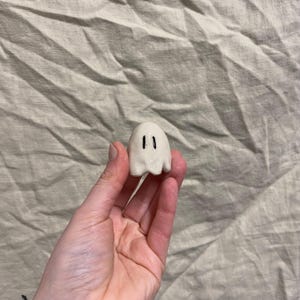 May include: A small, off-white ceramic ghost figurine with two black vertical eye markings. The ghost has a rounded body and a ruffled bottom edge. The figurine is held in a hand against a neutral-toned fabric background.