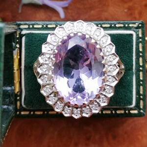 Op de afbeelding: Een ring met een grote ovale amethist-edelsteen, omringd door een halo van kleinere diamanten. De ring bevindt zich in een groene fluwelen ringdoos.