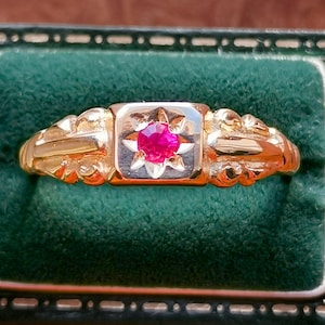 Op de afbeelding: Een gouden ring met een centrale vierkante zetting met een levendige rode edelsteen. De ring heeft sierlijke details op de band en wordt gepresenteerd in een groen fluwelen sieradendoosje. De edelsteen is omgeven door een stervormig ontwerp.