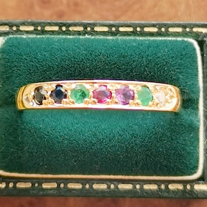 Puede incluir: Un anillo de oro con una fila de piedras preciosas de colores, incluyendo piedras negras, azules, verdes, rojas, rosas y verdes. El anillo se muestra en un joyero de terciopelo verde con un cierre dorado.