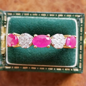 May include: A gold ring featuring three oval-cut vibrant pink rubies alternating with two circular clusters of small white diamonds. The ring is displayed horizontally, showcasing the five distinct gemstone settings set in a yellow gold band.