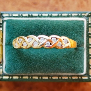 Peut inclure: Une bague en or présentant un motif métallique tressé ou entrelacé continu, sertie de six petites pierres rondes et transparentes. La bague est présentée horizontalement dans un écrin à bijoux doublé de velours vert foncé avec une bordure dorée.