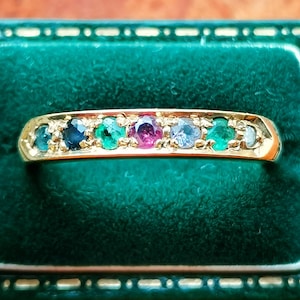 Puede incluir: Un anillo de oro con una fila de piedras preciosas de colores, incluyendo piedras azules, verdes y rosas. El anillo se muestra sobre un fondo de terciopelo verde, resaltando los colores brillantes de las piedras.