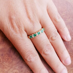 May include: A gold band ring featuring a row of five round green emeralds alternating with four small clear diamonds. The stones are set in a horizontal line across the top of the band, creating a classic multi-stone design on a polished gold setting.