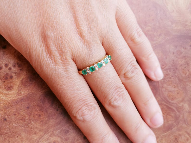 May include: A gold band ring featuring a row of five round green emeralds alternating with four small clear diamonds. The stones are set in a horizontal line across the top of the band, creating a classic multi-stone design on a polished gold setting.