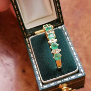 May include: A gold ring featuring a row of three round green emeralds alternating with pairs of small clear white diamonds, presented inside a dark green rectangular jewelry box with a cream-colored interior lining and gold-tone metal trim.