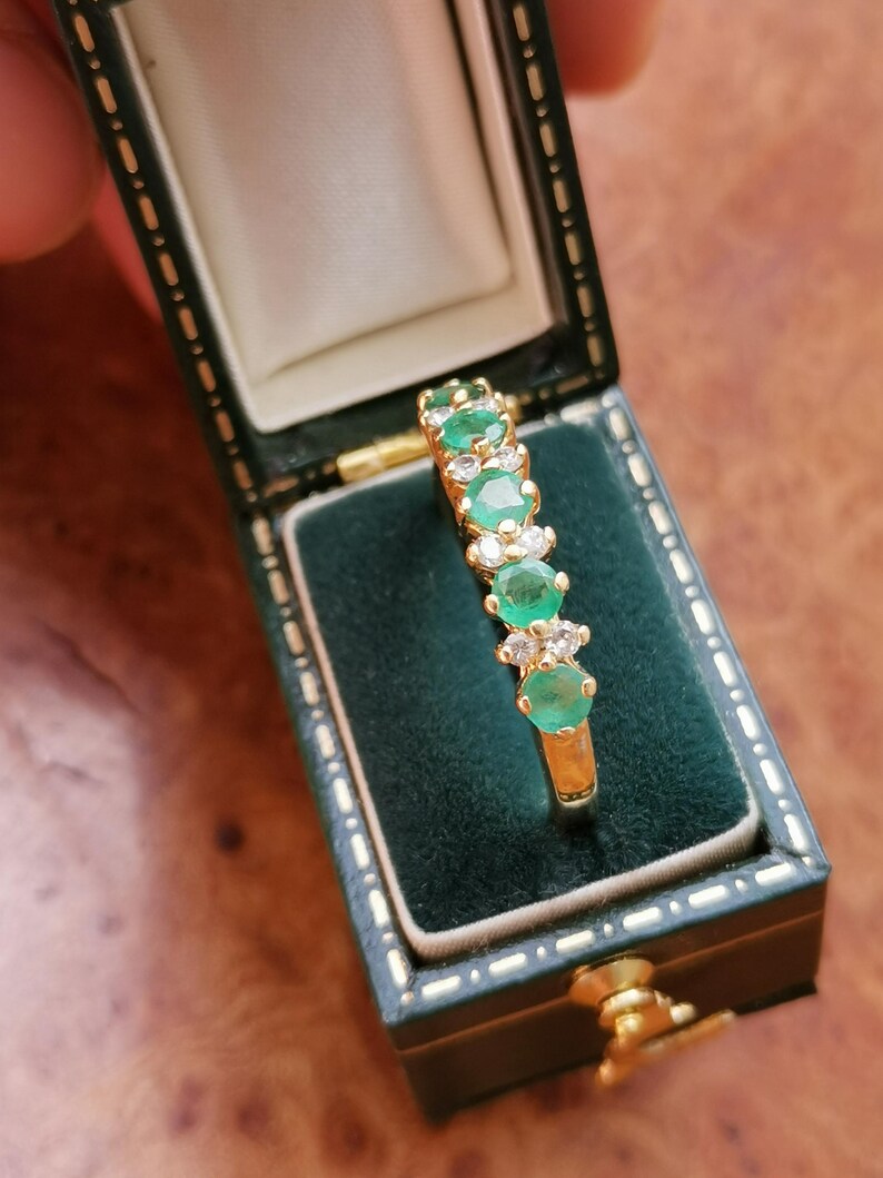 May include: A gold ring featuring a row of three round green emeralds alternating with pairs of small clear white diamonds, presented inside a dark green rectangular jewelry box with a cream-colored interior lining and gold-tone metal trim.