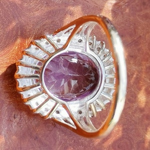 Op de afbeelding: Een zilveren ring met een grote ovale amethist-edelsteen omringd door kleinere diamanten. De ring heeft een delicate band.