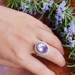 Op de afbeelding: Een zilveren ring met een grote ovale amethist-edelsteen omringd door kleinere diamanten. De ring is op een hand met een lichtbruine achtergrond.