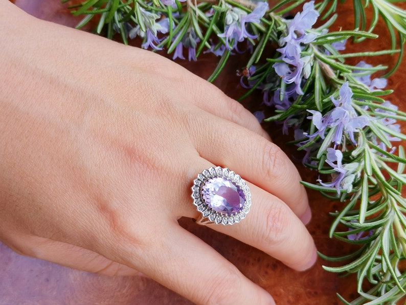 Op de afbeelding: Een zilveren ring met een grote ovale amethist-edelsteen omringd door kleinere diamanten. De ring is op een hand met een lichtbruine achtergrond.